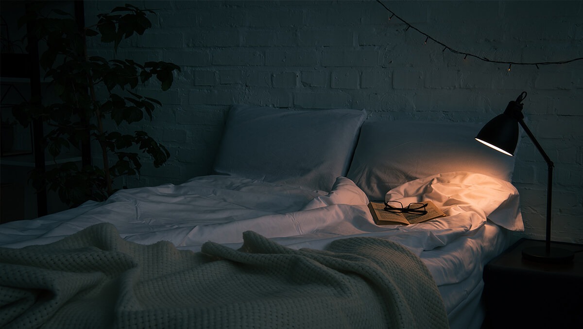Leeres Bett mit Buch und Lesebrille im Schein der Nachtischlampe