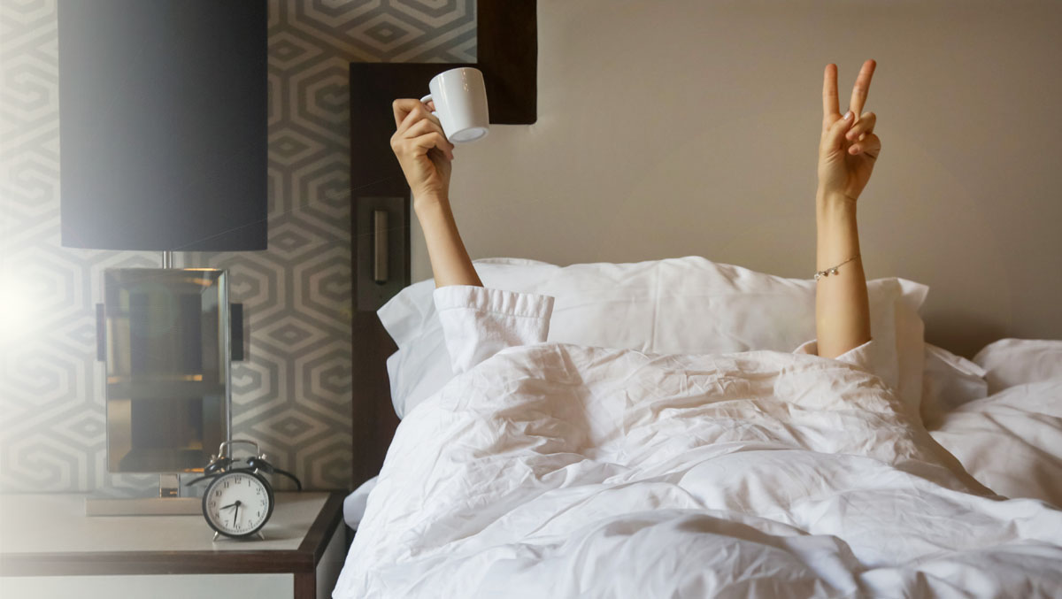 Frau im Bett hält Kaffeetasse in einer Hand und macht ein Peace-Zeichen