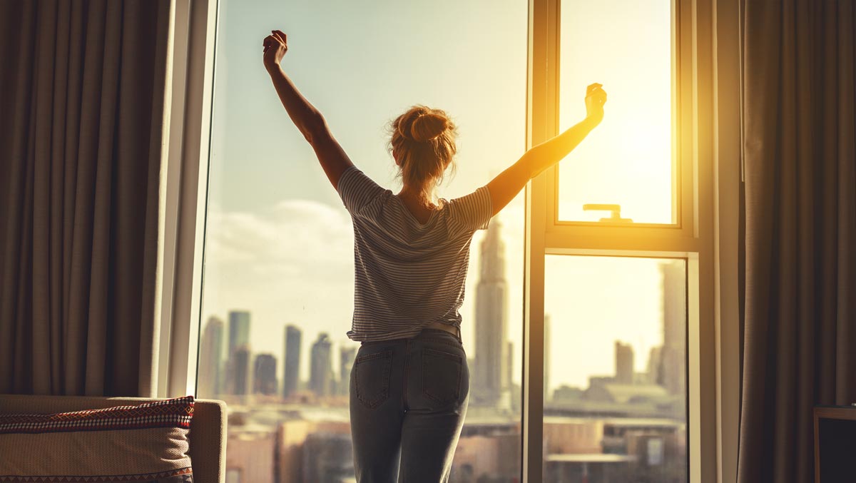 Morgens besser aus dem Bett kommen: Frau streckt sich vor Fenster bei Sonnenaufgang