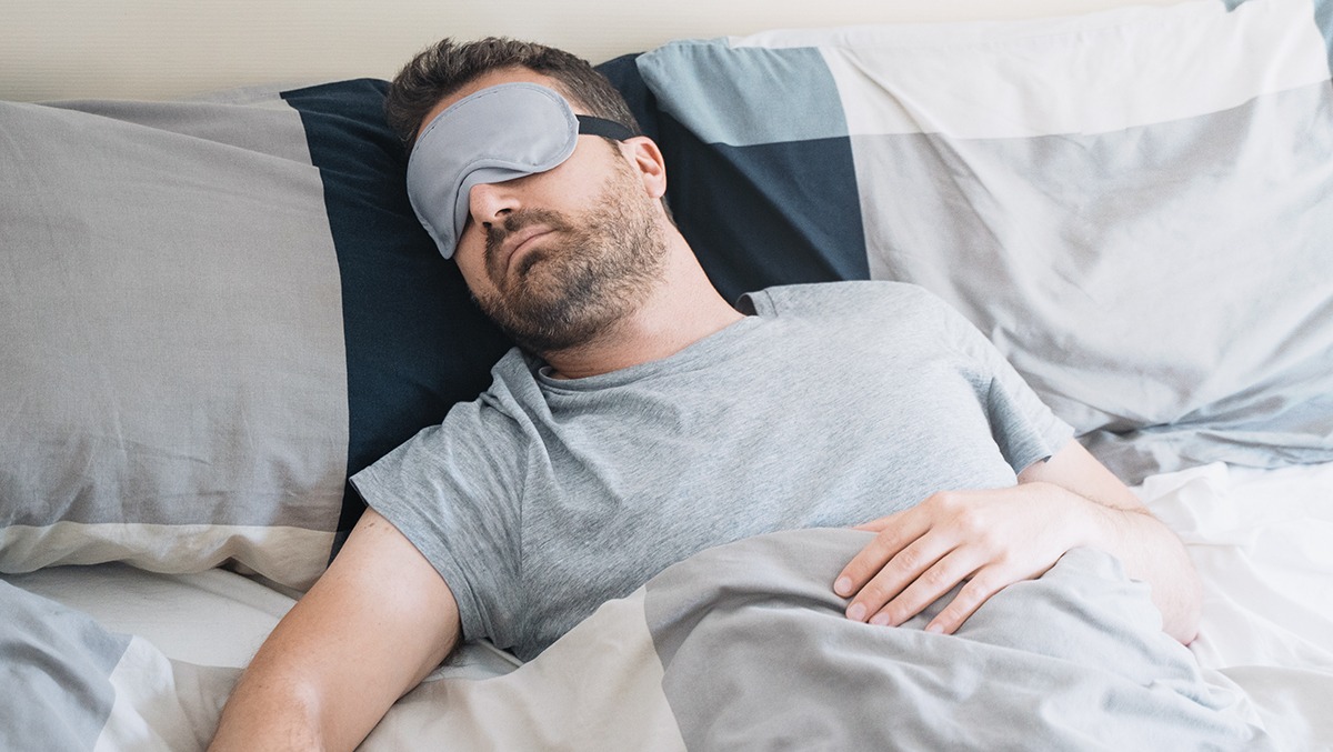 Mann liegt schlafend mit einer Augenmaske im Bett