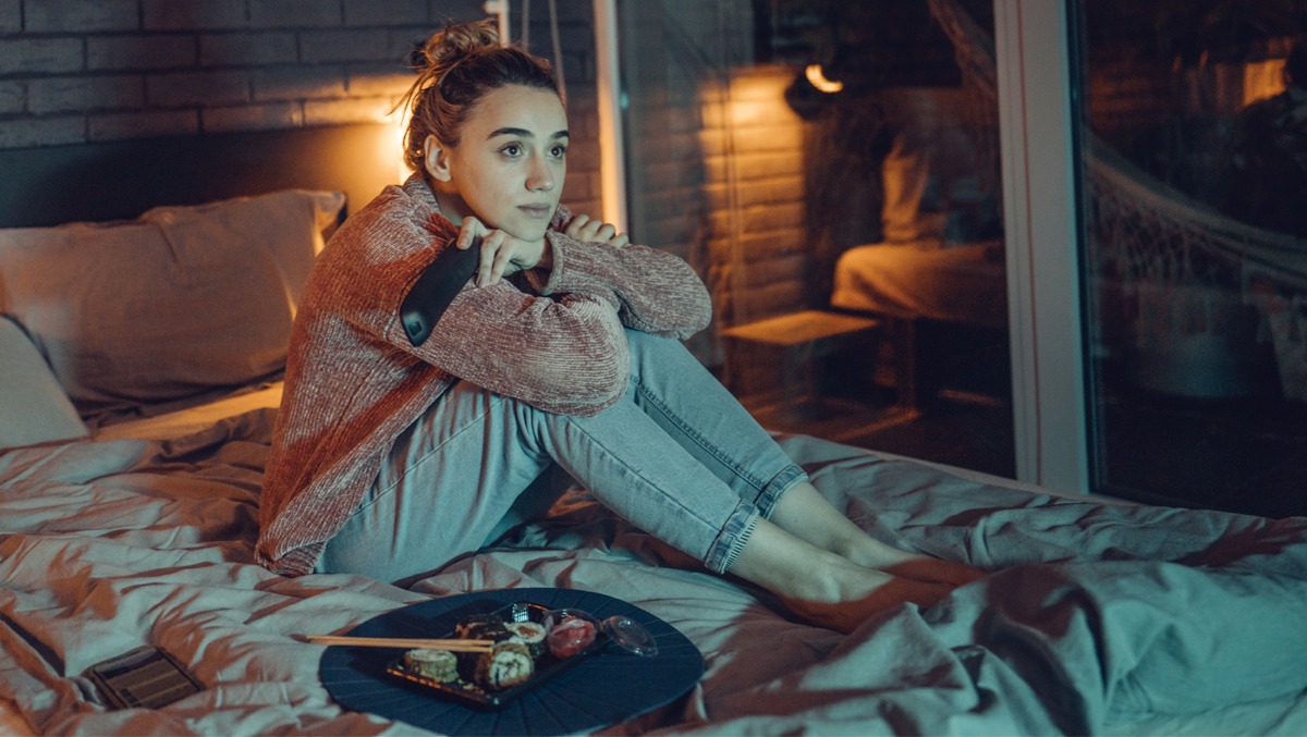 Junge Frau sitz auf dem Bett, neben ihr ein Teller Sushi
